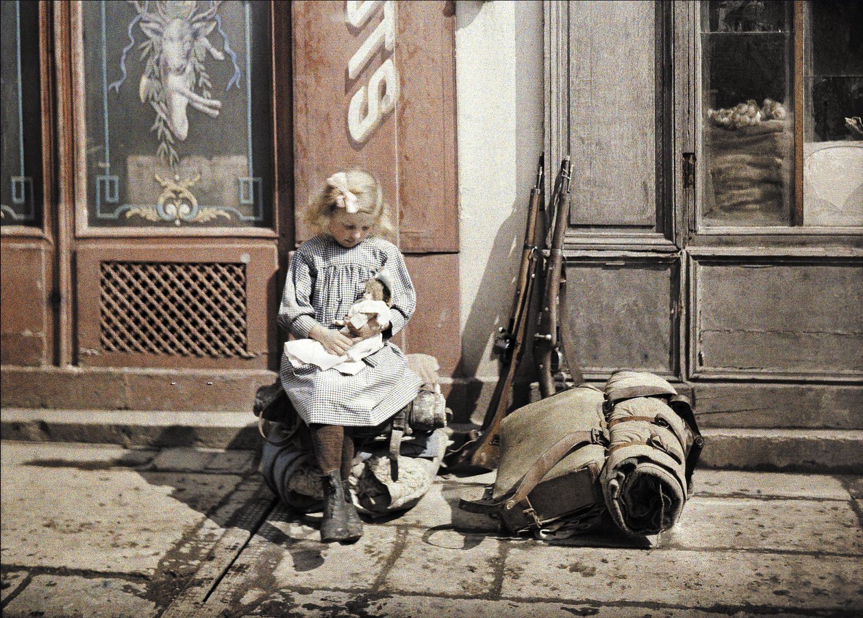 En lille pige leger med sin dukke; to pistoler og en rygsæk ligger ved siden af hende på jorden, Reims, Marne, Frankrig, 1917 (autokromt) af Fernand Cuville