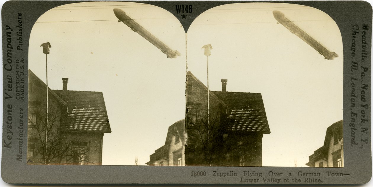 Horisontalt sepia -stereokort, der viser en Zeppelin, der flyver over tagene i en by. Keystone Stereograf nummer 18000. Titlen lyder af Keystone View Company