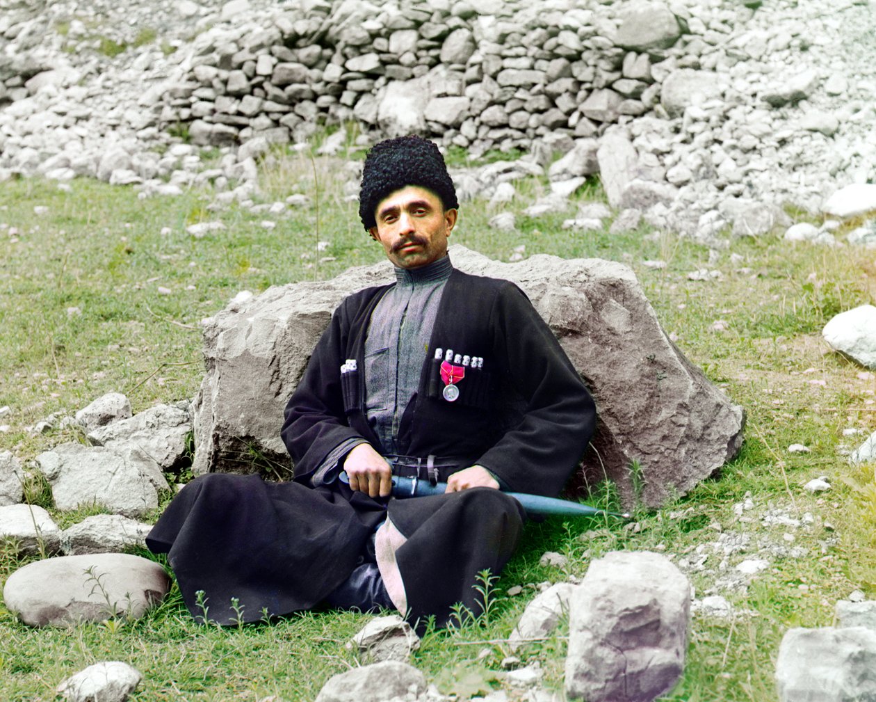 Dagestani-typer - Sunnimuslimsk mand iført traditionel kjole og hovedbeklædning med en beklædt dolk ved siden sidder på jorden, det russiske imperium, mellem 1905 og 1915 af Sergey Prokudin Gorsky