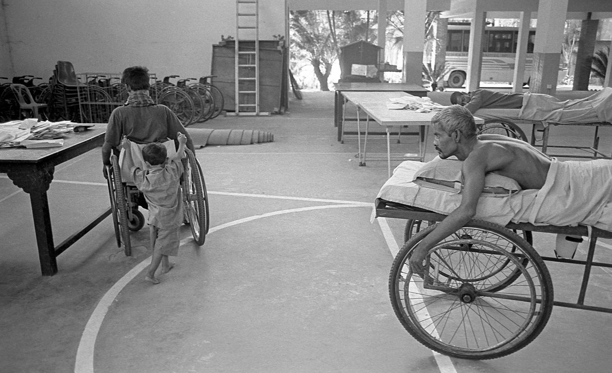 En fysisk udfordret mand i kørestol bliver båret af sin søn; en anden mand med samme situation ser på det i Center for Rehabilitering af Lammede, CRP i Savar, Dhaka, Bangladesh. 2005. af Unbekannt Unbekannt